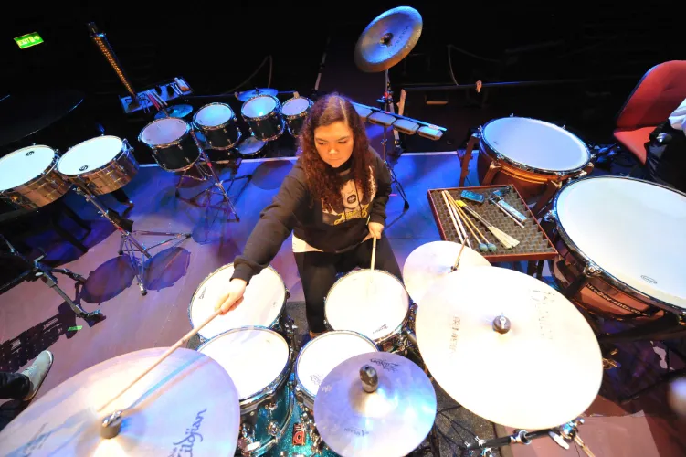 Female playing drum kit with concert toms, timbales and timpani in the background